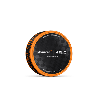 Lata redonda de color naranja y negro con las inscripciones «VELO» y «McLaren Spicy Papaya», decorada con un estampado a cuadros. En la tapa se lee «PEEL TO DISCOVER MORE» (Despecha para descubrir más). Esta edición limitada de VELO ofrece una experiencia única de nicotina con sabor a papaya afrutada y picante.