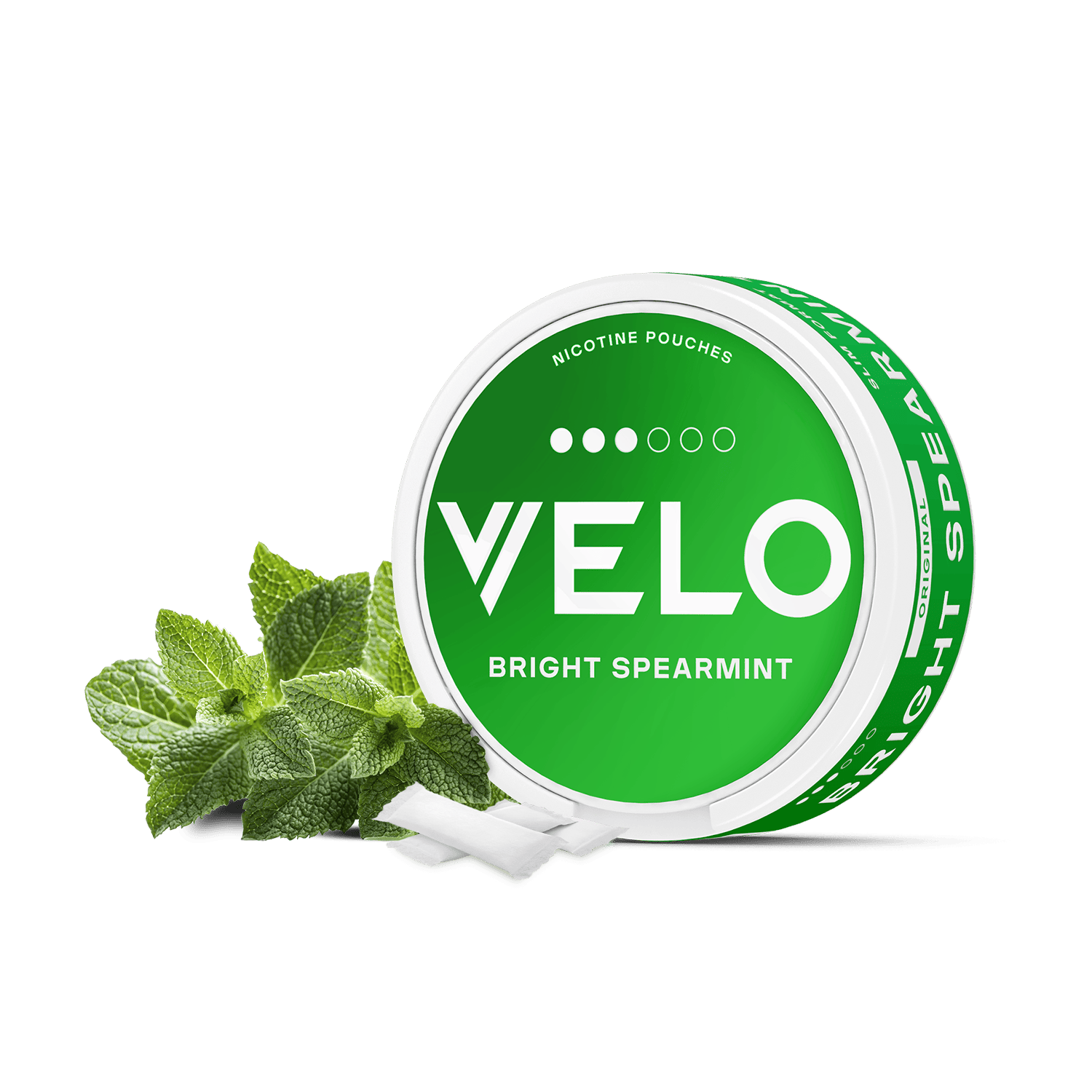 Un envase redondo verde y blanco de VELO Bright Spearmint se sitúa junto a hojas de menta fresca, con varias bolsitas blancas delante, destacando los ingredientes vegetales sobre un limpio fondo blanco.
