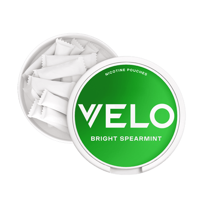 Envase redondo blanco de VELO Bright Spearmint con tapa verde, parcialmente abierto para mostrar varias bolsitas blancas en su interior. En la tapa aparece el texto "VELO BRIGHT SPEARMINT menta verde.