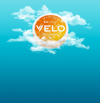 Un envase redondo naranja de VELO Tropical Ice aparece en el centro de un cielo azul con nubes blancas dispersas. El contenedor parece flotar entre las nubes.
