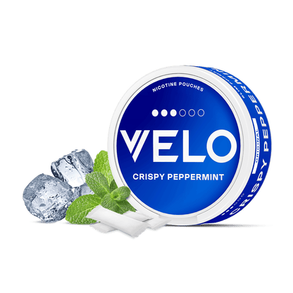 Un envase redondo azul y blanco de VELO Crispy Peppermint aparece junto a hojas de menta, chicles y cubitos de hielo, destacando su nicotina mentolada y sus ingredientes de alta calidad sobre un fondo blanco.
