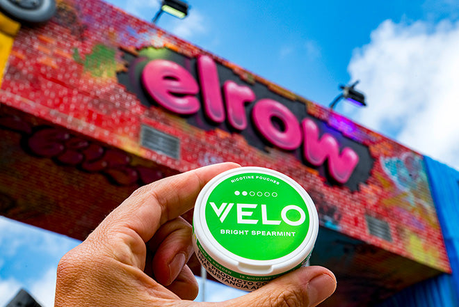 Una mano sostiene un envase de nicotina VELO Bright Spearmint verde y blanco delante de un colorido letrero de elrow sobre una estructura de ladrillo, con un cielo azul y nubes de fondo.
