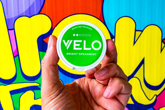 Una mano sostiene un envase redondo de bolsitas de nicotina VELO Bright Spearmint delante de un colorido fondo de estilo graffiti.