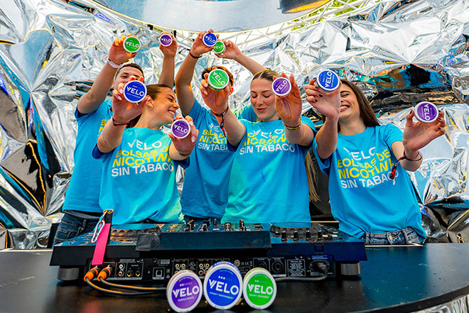 Cinco personas sonrientes con camisetas "VELO" azules a juego sostienen coloridos envases de bolsas de nicotina VELO detrás de una cabina de DJ, con brillantes decoraciones plateadas de fondo. También hay varios envases delante del equipo del DJ.