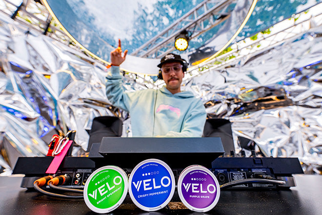 Un DJ con sudadera azul claro y auriculares pone música en una cabina metálica al aire libre. En primer plano, tres coloridos envases de nicotina VELO sobre una mesa.