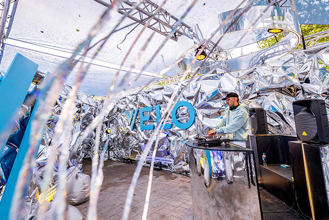 Un DJ actúa en una cabina para eventos al aire libre con decoraciones plateadas brillantes y un gran cartel VELO azul en el fondo metálico, bajo un toldo parcialmente cubierto.