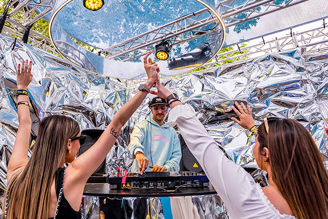Un DJ con sudadera azul claro actúa en un escenario metálico y brillante mientras dos mujeres en primer plano levantan las manos y bailan, disfrutando del animado ambiente al aire libre.