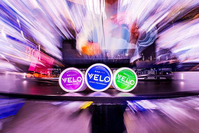 Tres envases redondos de nicotina VELO -morado, azul y verde- se muestran sobre una mesa con un fondo borroso y enérgico en el que aparece un DJ en un evento musical o una fiesta.