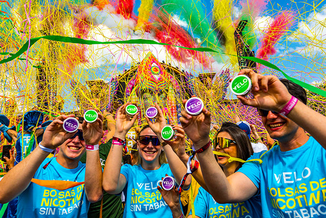 Un grupo de personas sonrientes con camisas azules sostienen coloridas latas circulares en un animado festival al aire libre, con serpentinas amarillas y rosas ondeando al fondo. El ambiente es alegre y festivo.