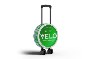 Una maleta redonda diseñada para parecerse a una lata verde de bolsitas de nicotina VELO Bright Spearmint, con asa y ruedas, en posición vertical sobre fondo blanco.