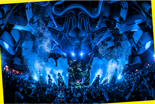 Una vibrante escena de concierto con una gran multitud vitoreando, luces azules, confeti y efectos de humo en el escenario, todo ello bajo un techo futurista y curvado. La enérgica atmósfera capta la emoción de un evento musical en directo.