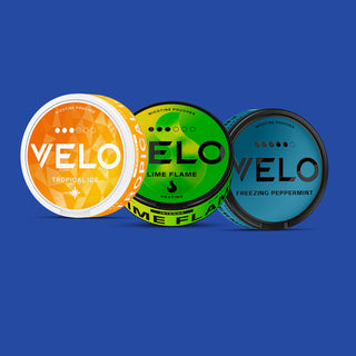 Tres latas redondas de bolsas de nicotina VELO se muestran una al lado de la otra sobre un fondo azul: Tropical Ice (naranja), Lime Flame (verde) y Freezing Peppermint (azul).