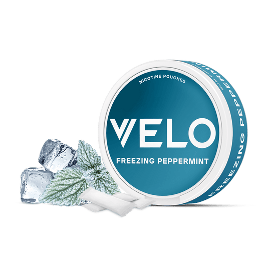 Aparece un envase redondo de VELO Freezing Peppermint con cubitos de hielo, una hoja de menta escarchada y dos bolsitas blancas sobre un fondo blanco, mostrando su intenso sabor a menta y sus ingredientes de alta calidad.