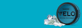Se muestra una lata redonda de bolsitas de nicotina VELO Freezing Peppermint con cubitos de hielo, una hoja de menta y una rama de pino sobre un fondo azul brillante.