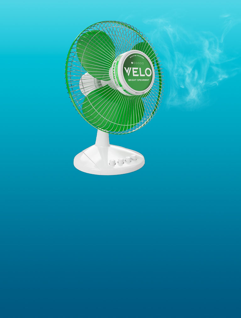 Un ventilador de mesa eléctrico verde y blanco con la palabra VELO en el centro, que sopla niebla o vapor visible sobre un fondo azul degradado.