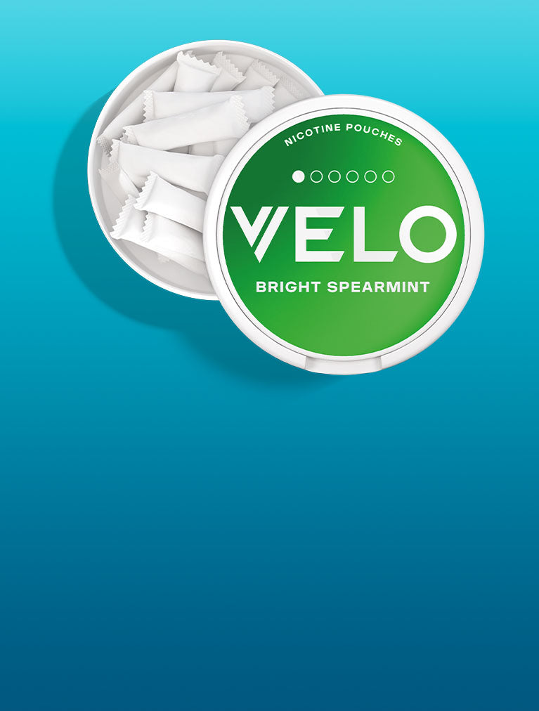 Un recipiente redondo y blanco con la etiqueta VELO Bright Spearmint contiene varias bolsitas de nicotina blancas y rectangulares sobre un fondo azul degradado. La tapa del envase está parcialmente abierta, dejando al descubierto las bolsitas que contiene.