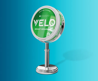 Una lata redonda de bolsitas de nicotina VELO Bright Spearmint se exhibe en un brillante soporte plateado sobre un fondo azul degradado.