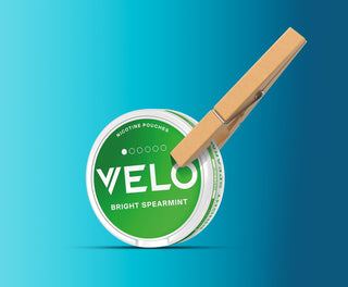 Un envase redondo de nicotina VELO Bright Spearmint se sujeta con una gran pinza de madera sobre un fondo azul degradado.