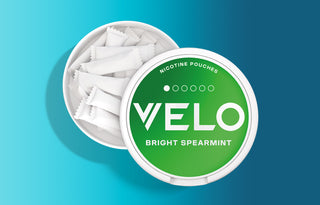 Un envase redondo de sobres de nicotina VELO Bright Spearmint con la tapa parcialmente abierta, mostrando varios sobres blancos en su interior. La tapa es verde con texto blanco sobre fondo azul degradado.