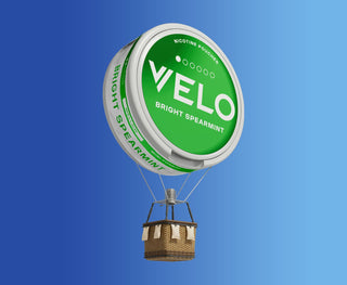 Un globo aerostático con una bolsa de nicotina VELO Bright Spearmint verde y blanca como globo y una cesta de mimbre debajo, flotando sobre un fondo azul degradado.