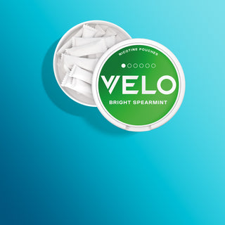Sobre un fondo azul degradado se muestra un envase blanco redondo de sobres de nicotina VELO Bright Spearmint, abierto con varios sobres blancos en su interior.