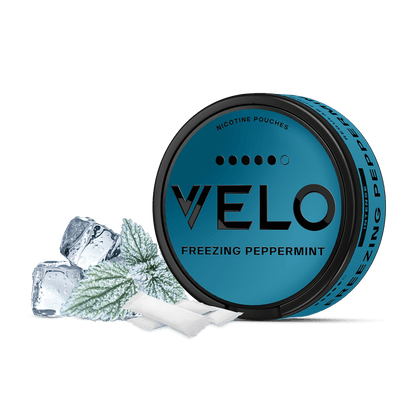 Una lata redonda de color azul con la etiqueta VELO Freezing Peppermint se encuentra junto a cubitos de hielo, una hoja de menta y dos bolsitas sobre un fondo blanco, resaltando su intenso sabor a menta y su nicotina mentolada de alta calidad.