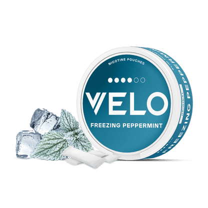 Un envase redondo de VELO Freezing Peppermint se representa con cubitos de hielo, una hoja de menta y dos bolsitas blancas delante. El envase azul y blanco presenta ingredientes de alta calidad e intensa menta para un fresco sabor a menta.