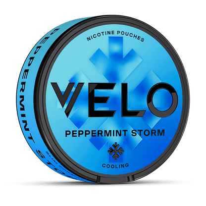 Una lata VELO redonda de color azul etiquetada como Peppermint Storm presenta un texto en negrita de color negro, un gráfico de cristales de hielo y "Cooling" más "sabor a menta intensa" en la tapa. El lateral destaca ingredientes de origen vegetal y el nombre del producto.