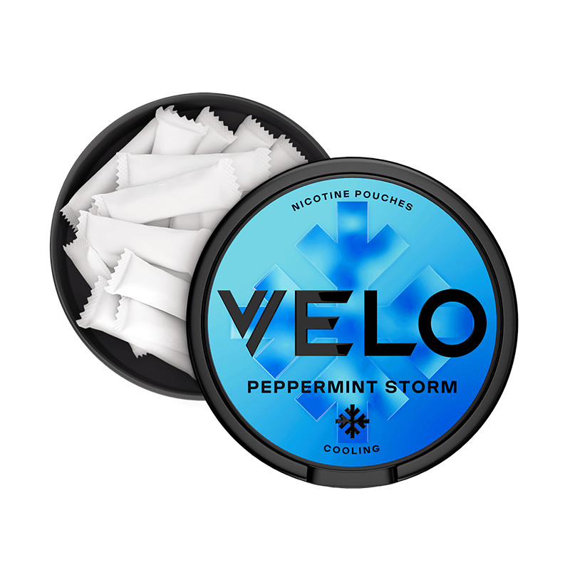 Un envase VELO negro, redondo y abierto, con una tapa azul etiquetada "Peppermint Storm", muestra bolsitas blancas en su interior. La tapa muestra un copo de nieve y la palabra "cooling" (enfriamiento), destacando su intenso sabor a menta y su nicotina mentolada.
