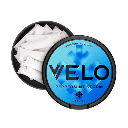 Un envase VELO negro, redondo y abierto, con una tapa azul etiquetada "Peppermint Storm", muestra bolsitas blancas en su interior. La tapa muestra un copo de nieve y la palabra "cooling" (enfriamiento), destacando su intenso sabor a menta y su nicotina mentolada.