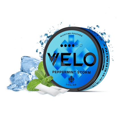 Un envase redondo azul de VELO Peppermint Storm se sitúa junto a cubitos de hielo, hojas de menta y dos bolsitas blancas, destacando su intenso sabor a menta y su nicotina de origen vegetal.