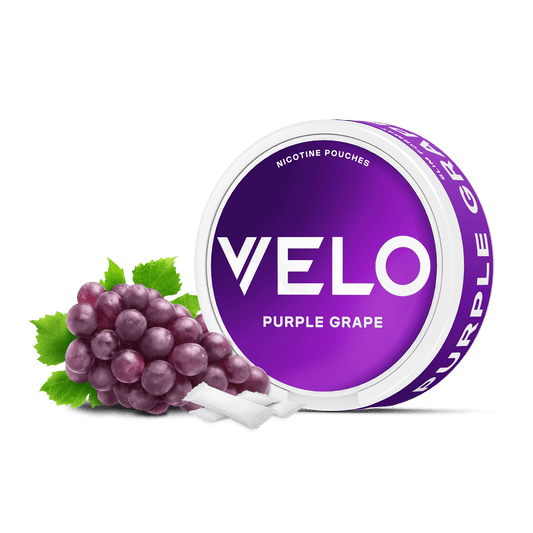 Una lata redonda de color púrpura con la etiqueta VELO Purple Grape y texto en blanco se sitúa junto a un racimo de uvas dulces y varias bolsitas blancas de nicotina, destacando su sabor afrutado a nicotina, sobre un fondo blanco.