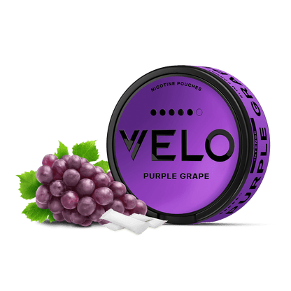 Junto a un racimo de uvas y dos bolsitas de nicotina blancas sobre fondo blanco, hay una lata redonda de color morado con la etiqueta VELO Purple Grape.