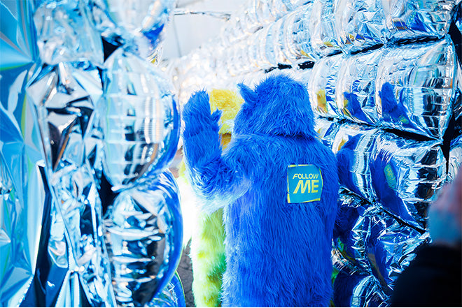 Dos personas con disfraces peludos de monstruos azules y amarillos caminan por un túnel bordeado de globos plateados brillantes. El disfraz azul tiene un cartel en la espalda que dice FOLLOW ME.