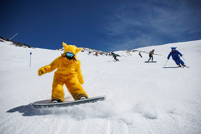 Una persona con un disfraz de animal amarillo brillante practica snowboard por una pendiente nevada, seguida de otras personas con disfraces y equipo de esquí, bajo un cielo azul despejado.