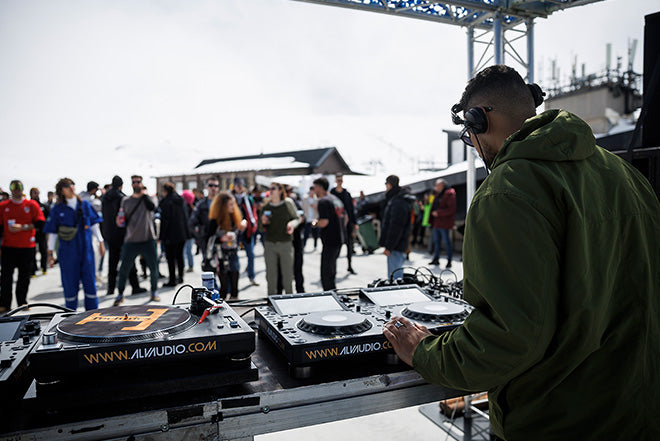 Un DJ con auriculares toca música en un tocadiscos en un evento al aire libre, con una multitud de personas reunidas en el fondo bajo un cielo nublado.