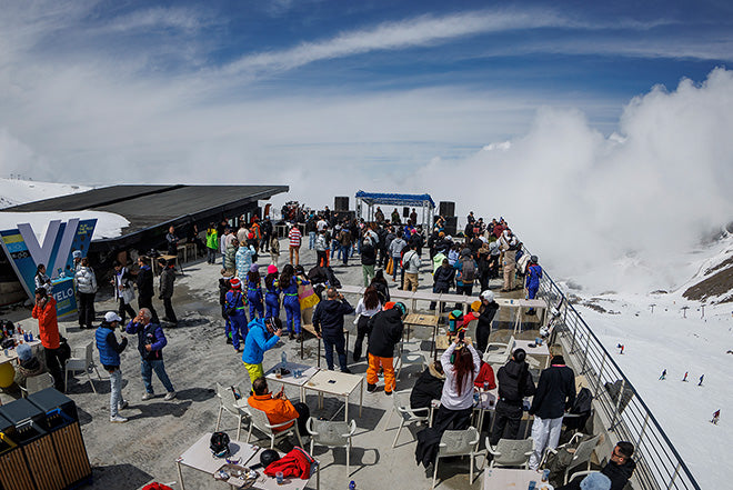 Una animada terraza al aire libre en una montaña nevada, abarrotada de gente con coloridos trajes de esquí comiendo, bebiendo y socializando bajo un cielo parcialmente nublado, con una ladera cubierta de nieve y picos lejanos al fondo.