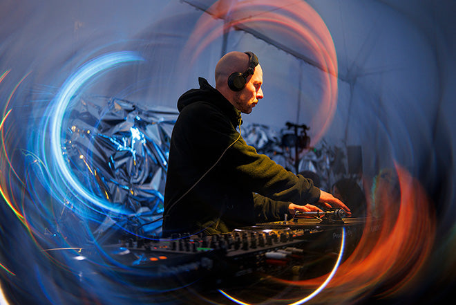 Un DJ con auriculares y sudadera oscura actúa ante una mesa de mezclas, rodeado de remolinos de luces de colores en un local decorado en tonos metálicos.