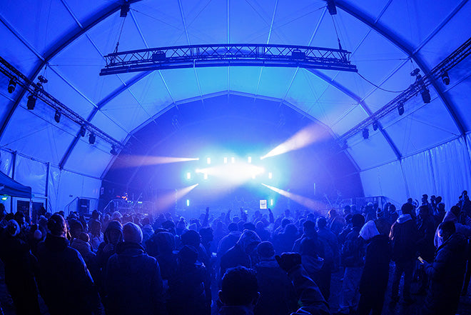 Una gran multitud se reúne de noche en el interior de una carpa, frente a un escenario brillantemente iluminado con focos azules y morados que brillan hacia el público. El ambiente parece animado y enérgico.