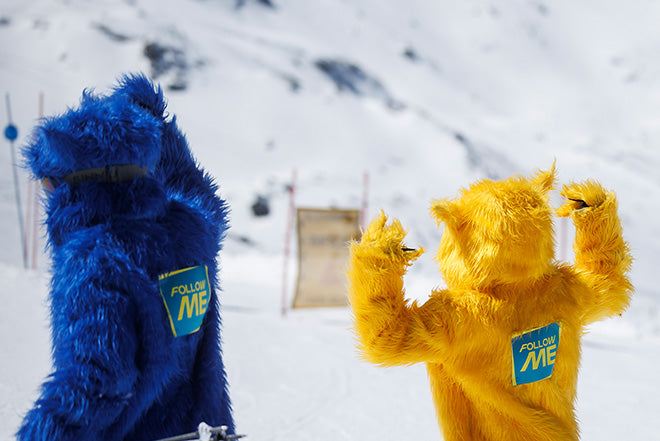 Dos personas vestidas con trajes de peluche azules y amarillos y con carteles de "FOLLOW ME" a la espalda están de pie en una ladera nevada, de espaldas a la cámara, con bastones de esquí en la mano y montañas al fondo.