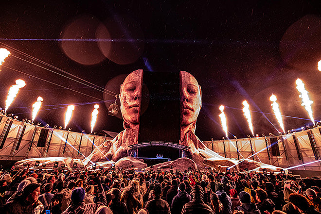 Una gran multitud se reúne en un festival de música al aire libre frente a un espectacular escenario con dos gigantescos rostros esculpidos y columnas de fuego que se disparan hacia el cielo nocturno.
