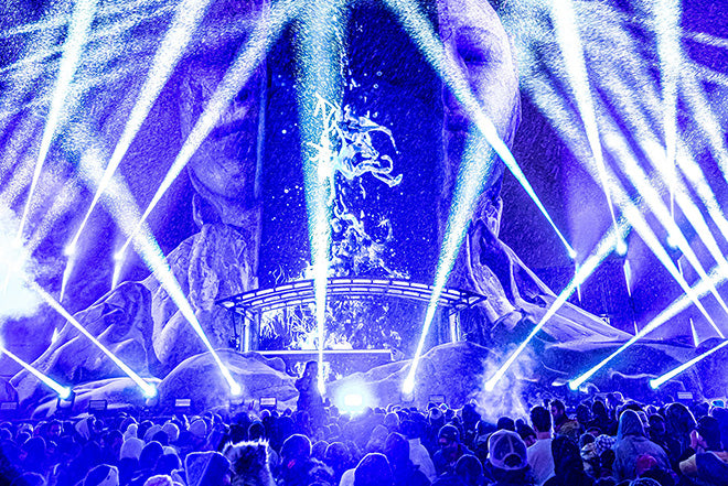 Una vibrante escena de concierto con una multitud frente a un escenario brillantemente iluminado, con espectaculares focos azules y blancos y efectos similares a la nieve, con grandes figuras esculpidas como telón de fondo del escenario.
