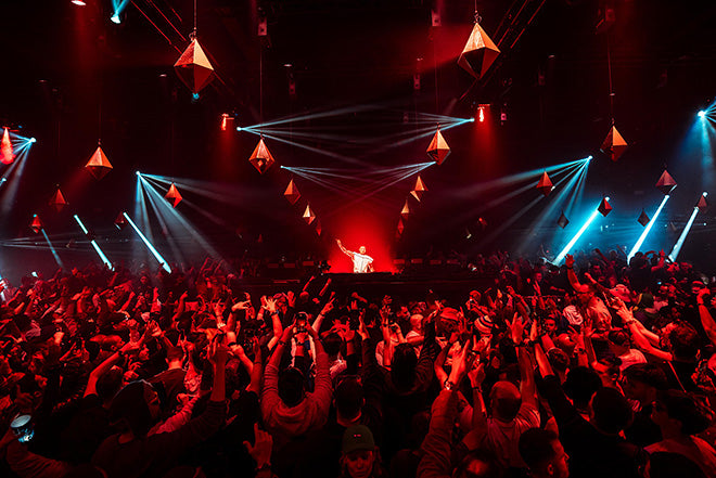Una multitud baila y anima frente a un DJ en un escenario, iluminado por luces rojas y azules y adornos geométricos colgantes en una oscura sala de conciertos.