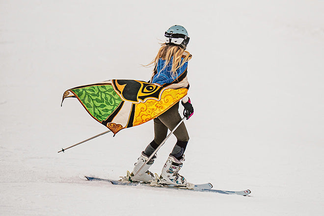 Un esquiador con una capa y un casco de colores esquía en una pista nevada, visto desde atrás. La capa presenta motivos verdes, amarillos, negros y naranjas que recuerdan a las alas de un pájaro o un dragón.
