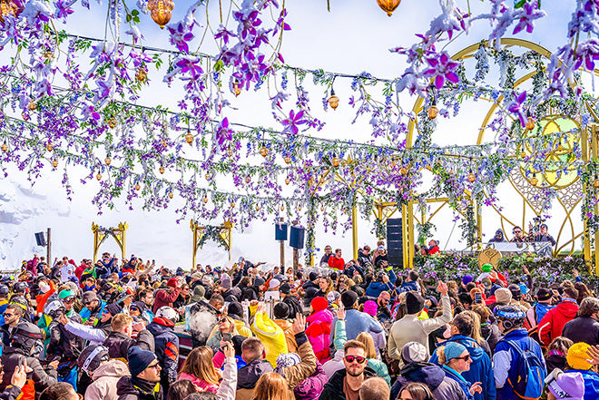 Una gran multitud disfruta de un festival de música al aire libre rodeado de nieve, con vibrantes flores púrpuras y vegetación colgando por encima y DJs tocando en un escenario decorado al fondo.