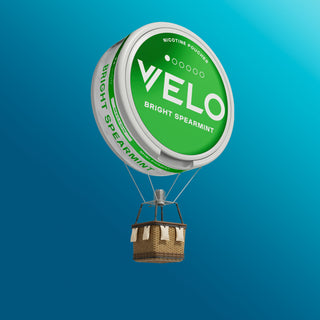Un globo aerostático con una cesta de mimbre está coronado por un gran envase de nicotina VELO Bright Spearmint verde y blanco, flotando sobre un fondo azul.