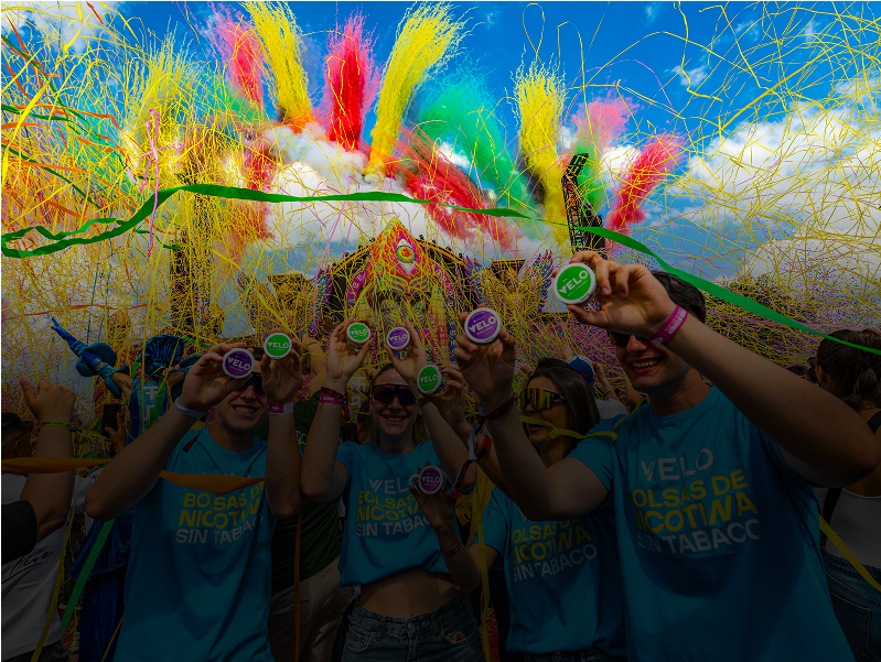 Un grupo de personas en un colorido festival al aire libre sostiene insignias redondas. El cielo se llena de serpentinas brillantes y confeti, y los participantes llevan camisetas azules con texto amarillo que promueven un estilo de vida sin nicotina.