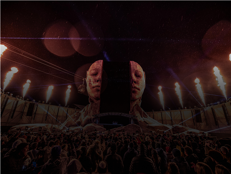 Una gran multitud se enfrenta a un escenario dramático en el que aparecen dos rostros humanos gigantes iluminados con luces ardientes y llamas que salen disparadas hacia arriba por la noche. La escena es atmosférica e intensa, con cielos oscuros por encima.